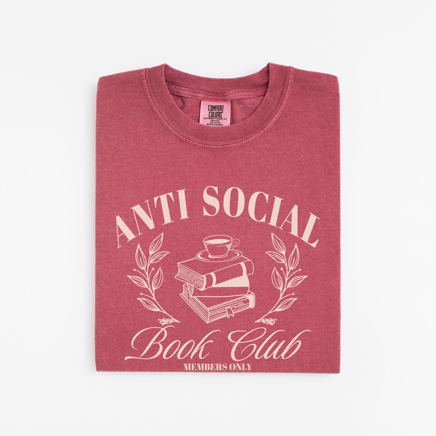 A folded unisex Comfort Colors Shirt in the color crimson. The design on the shirt features 'Anti Social Book Club - Members Only' text and book stack design in cream color