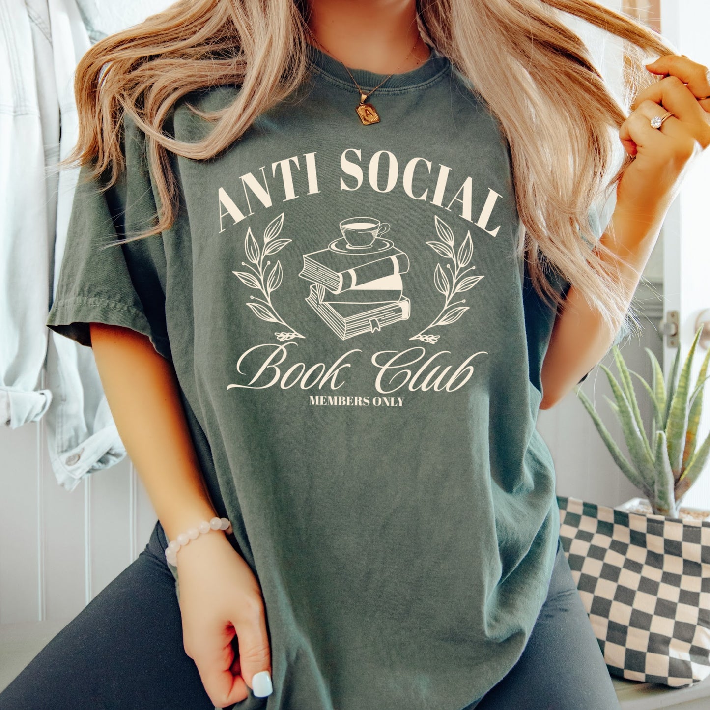 A woman wearing a unisex Comfort Colors Shirt in the color moss.The design on the shirt features 'Anti Social Book Club - Members Only' text and book stack design in cream color