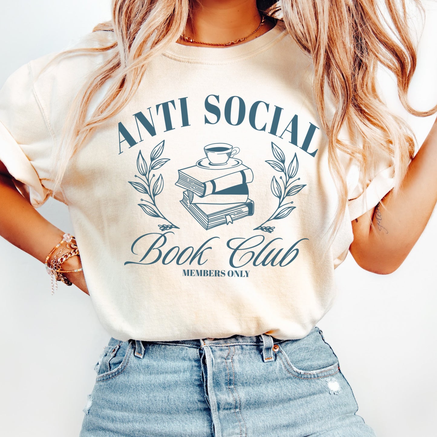 A woman wearing a unisex Comfort Colors Shirt in the color ivory. The design on the shirt features 'Anti Social Book Club - Members Only' text and book stack design in dark teal color