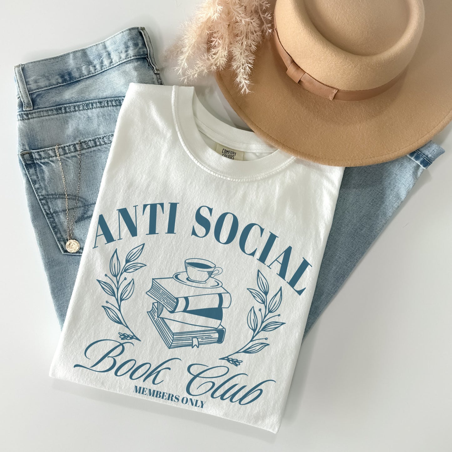 A folded unisex Comfort Colors Shirt in the color white.The design on the shirt features 'Anti Social Book Club - Members Only' text and book stack design in dark teal color