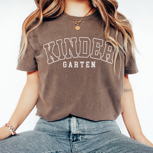 A woman wearing a unisex Comfort Colors t-shirt in espresso color with 'Kindergarten' printed in bold varsity-style lettering.