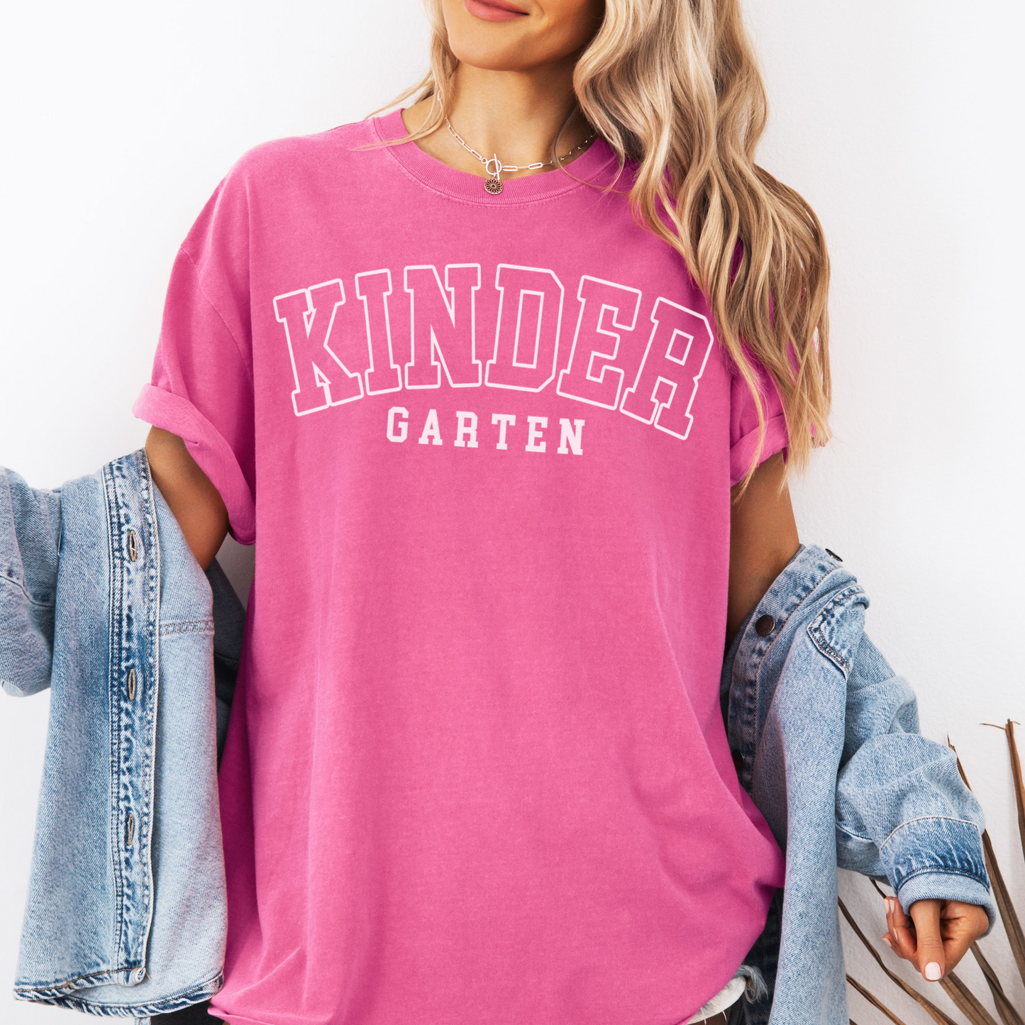 A woman wearing a unisex Comfort Colors t-shirt in crunchberry color with 'Kindergarten' printed in bold varsity-style lettering.