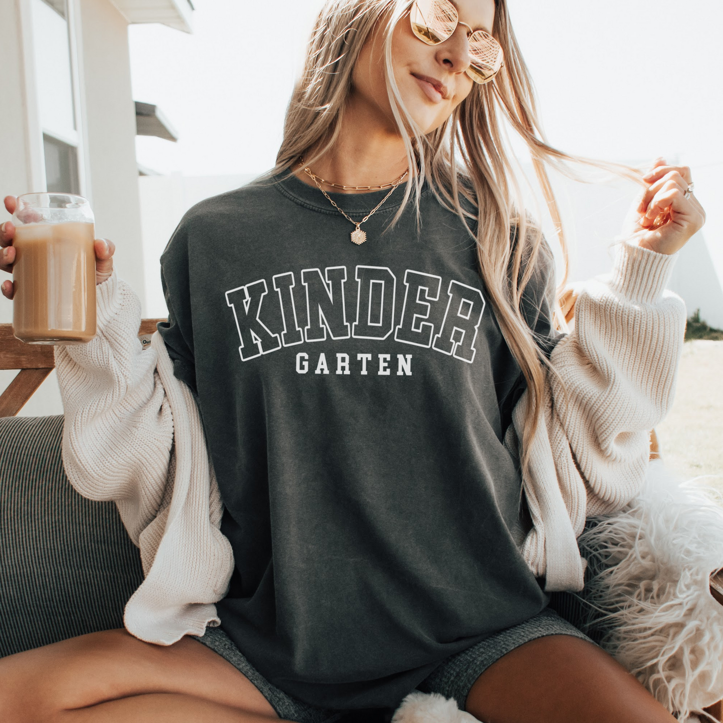 A woman wearing a unisex Comfort Colors t-shirt in pepper color with 'Kindergarten' printed in bold varsity-style lettering.