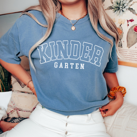 A woman wearing a unisex Comfort Colors t-shirt in blue jean color with 'Kindergarten' printed in bold varsity-style lettering.