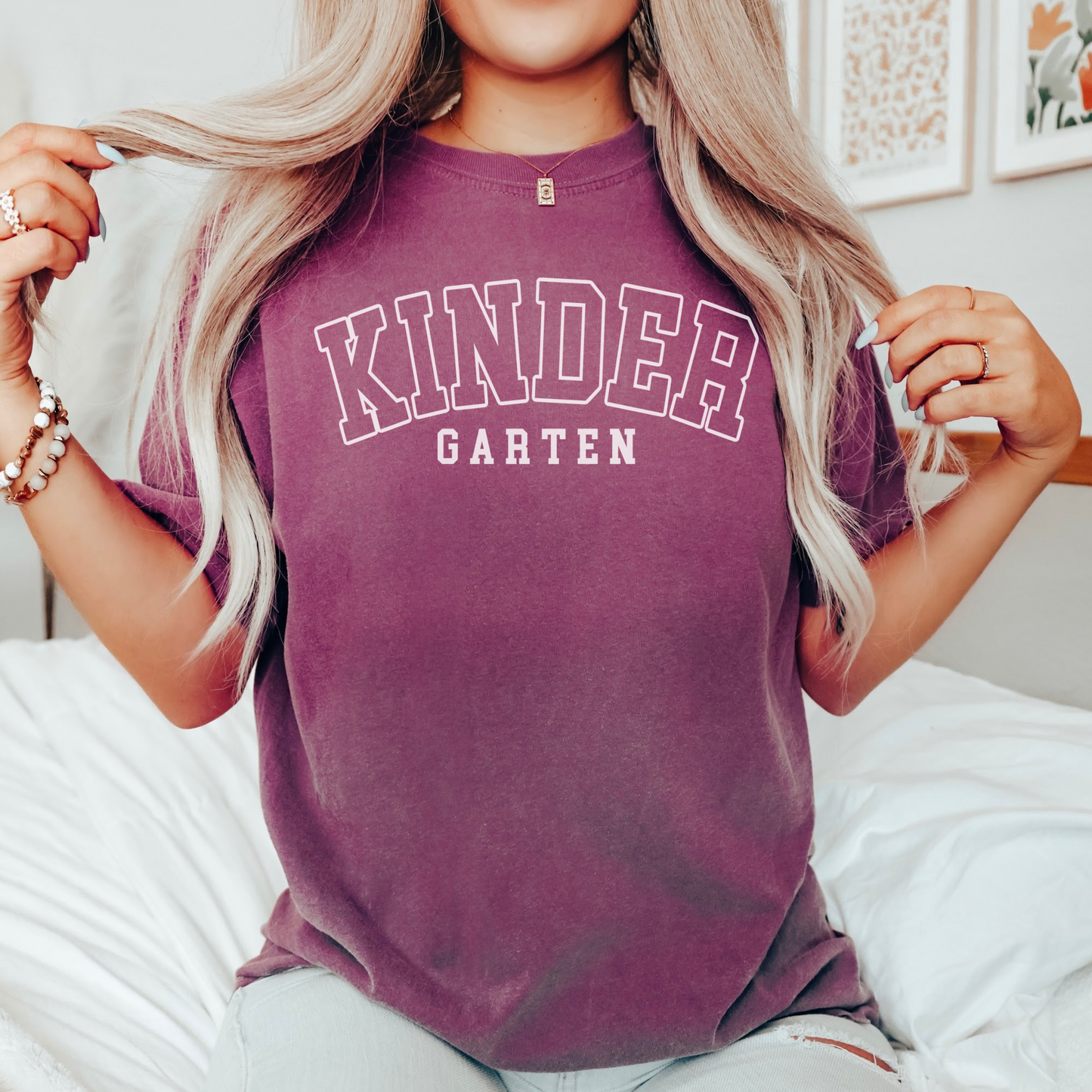 A woman wearing a unisex Comfort Colors t-shirt in berry color with 'Kindergarten' printed in bold varsity-style lettering.