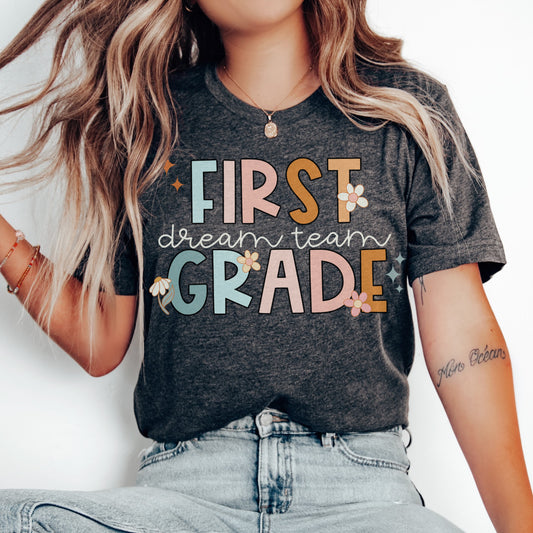 "First Grade Dream Team" with bright, multicolored font and cursive lettering. Random star and flower elements places around design.Unisex women's Tshirt, color: dark grey heather