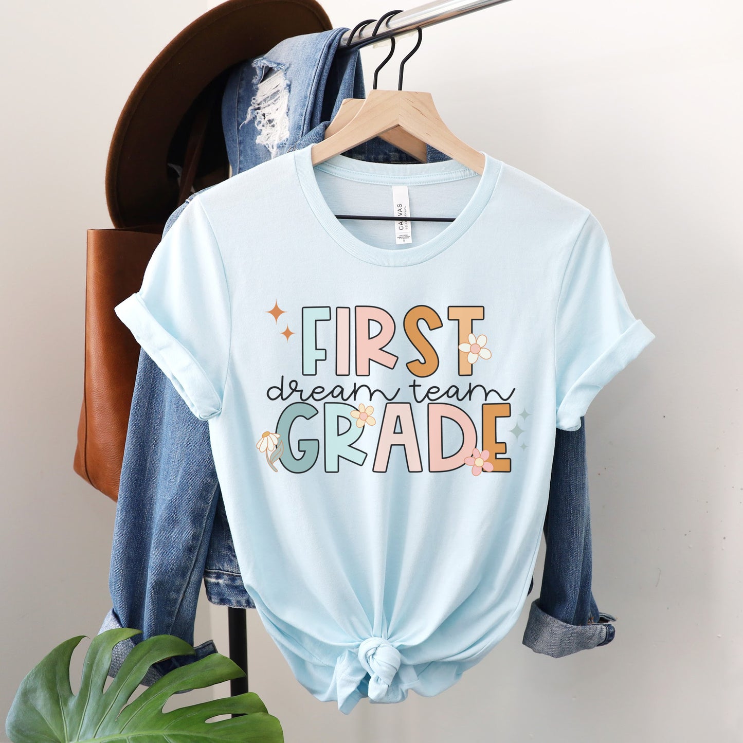 "First Grade Dream Team" with bright, multicolored font and cursive lettering. Random star and flower elements places around design.Unisex women's Tshirt, color: Heather Ice Blue