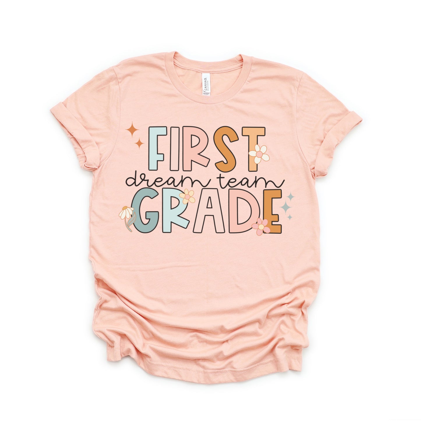 "First Grade Dream Team" with bright, multicolored font and cursive lettering. Random star and flower elements places around design.Unisex women's Tshirt, color: Heather Peach