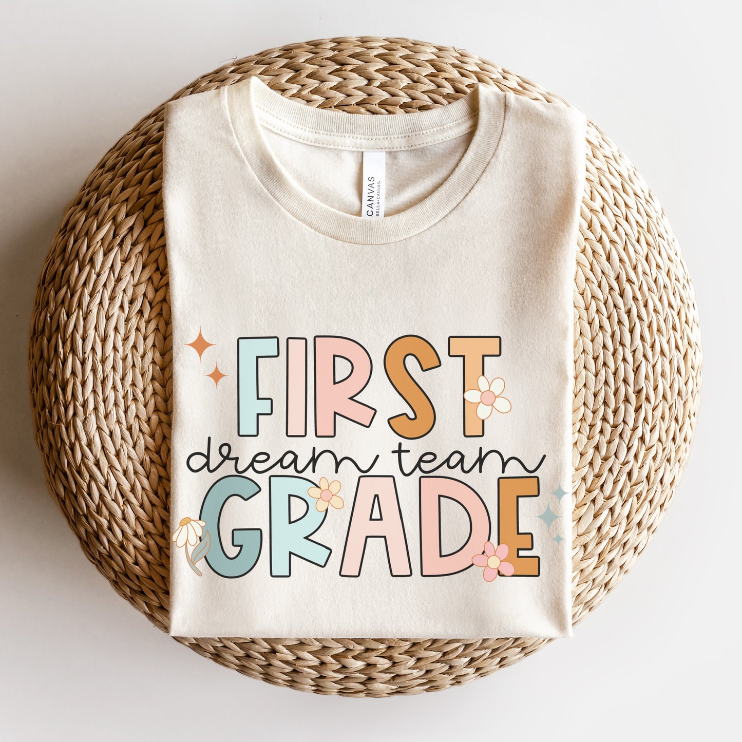 "First Grade Dream Team" with bright, multicolored font and cursive lettering. Random star and flower elements places around design.Unisex women's Tshirt, color: Natural