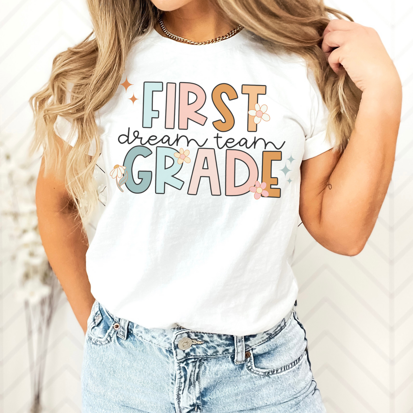 "First Grade Dream Team" with bright, multicolored font and cursive lettering. Random star and flower elements places around design.Unisex women's Tshirt, color: White