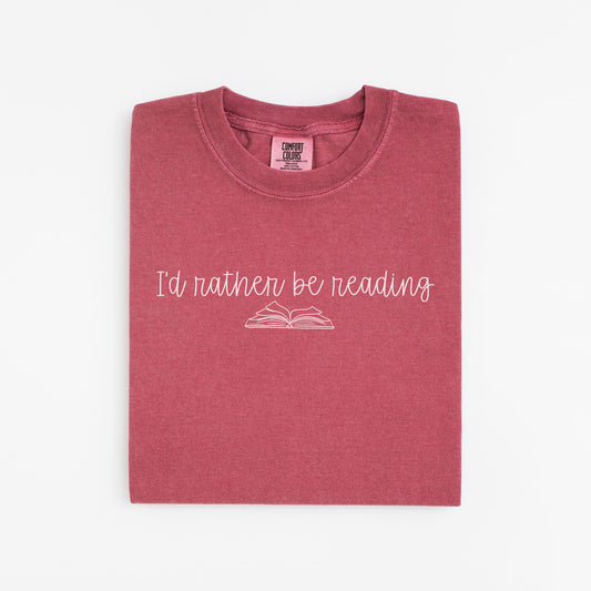 A folded unisex Comfort Colors t-shirt in crimson color with the phrase 'I'd rather be reading' printed above a simple line drawing of an open book. 