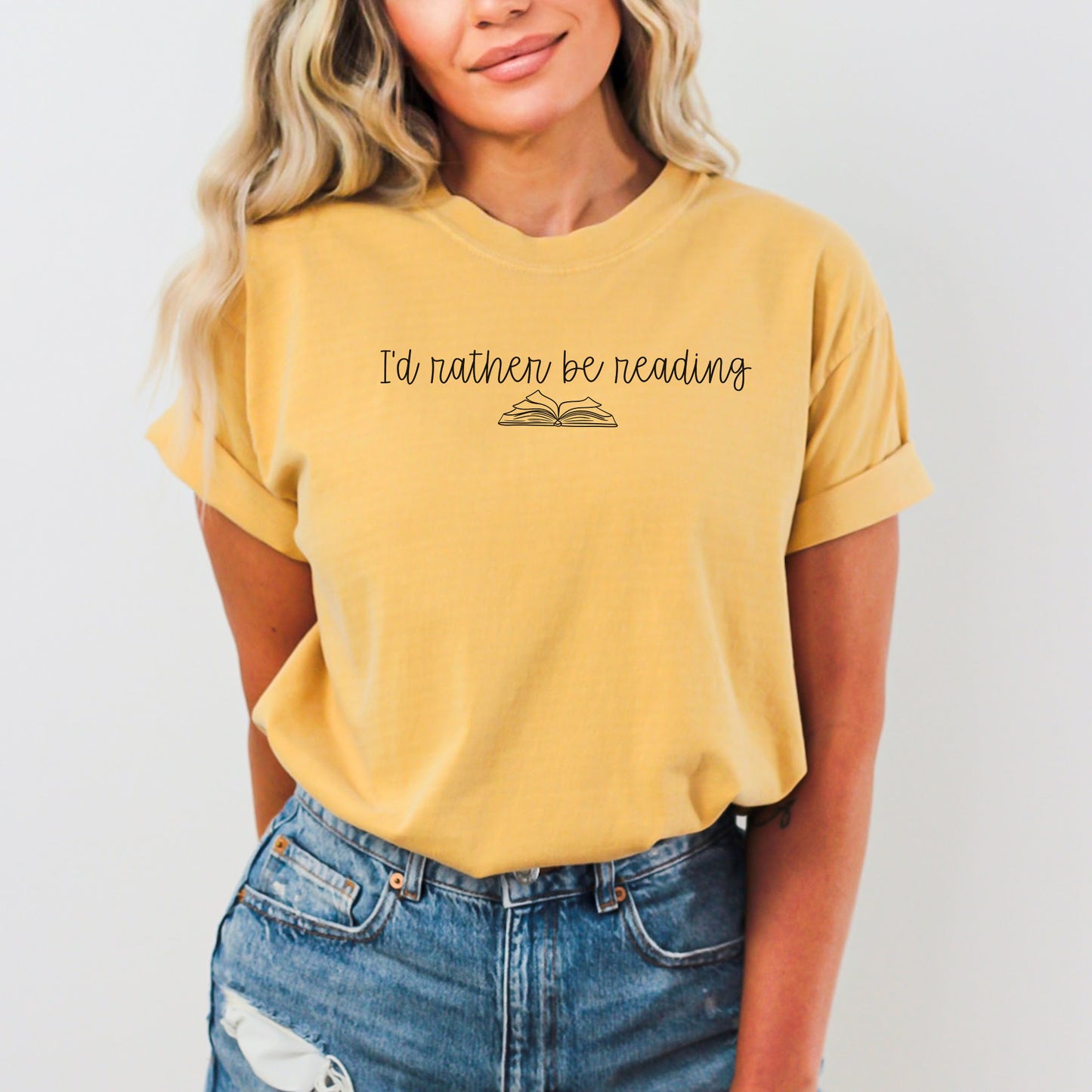 A woman wearing a unisex Comfort Colors t-shirt in mustard color with the phrase 'I'd rather be reading' printed above a simple line drawing of an open book.