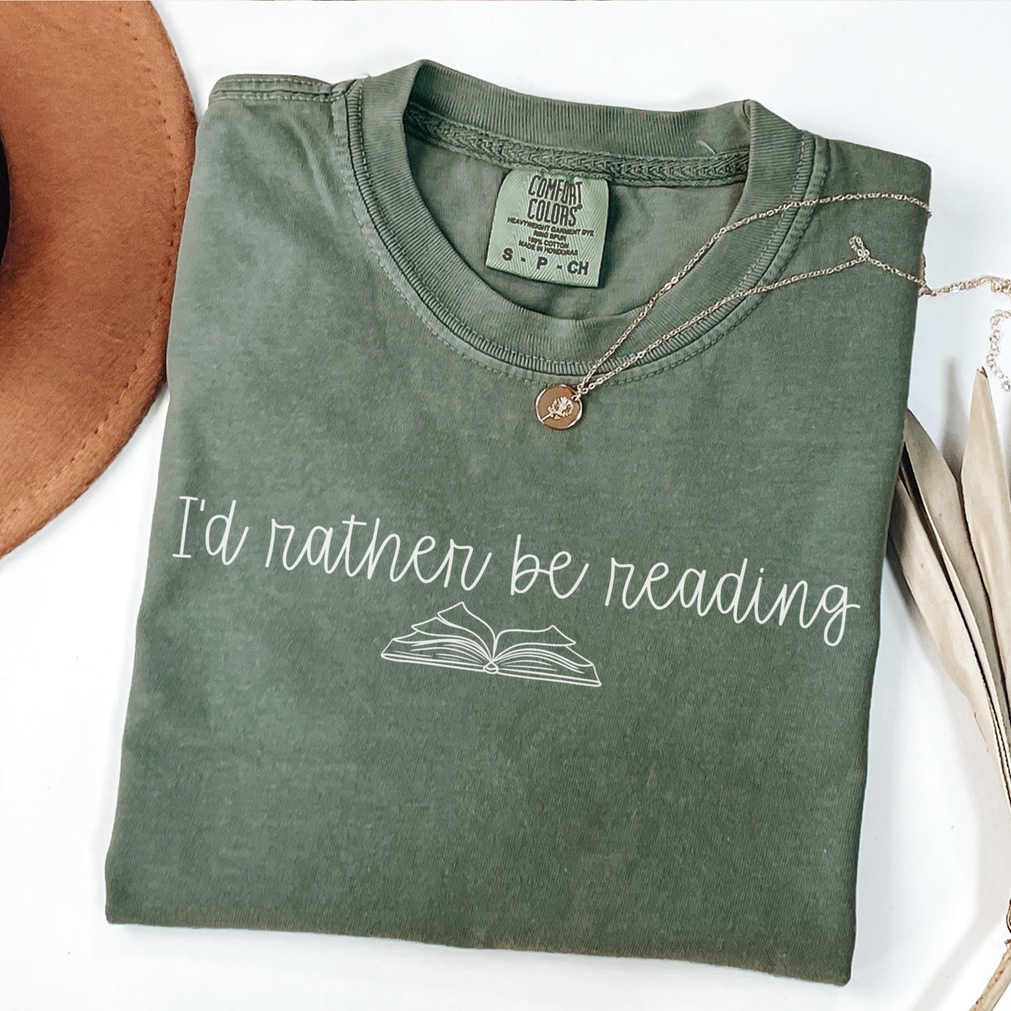 A folded unisex Comfort Colors t-shirt in moss color, displayed with a necklace and a hat beside it. The shirt features the phrase 'I'd rather be reading' printed above a simple line drawing of an open book.