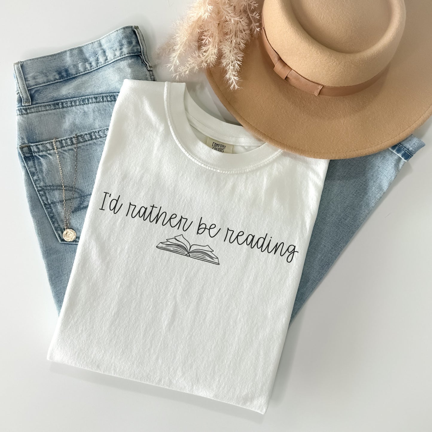 A folded unisex Comfort Colors t-shirt in white, displayed with a pair of light blue jeans, a tan hat, and a necklace. The shirt features the phrase 'I'd rather be reading' printed above a simple line drawing of an open book.