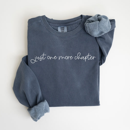 A folded unisex Comfort Colors sweatshirt in denim color, the phrase 'Just one more chapter' in elegant white script across the chest