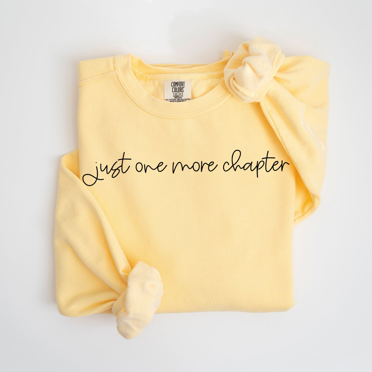 A folded unisex Comfort Colors sweatshirt in butter color, featuring the phrase 'Just one more chapter' in elegant black script across the chest