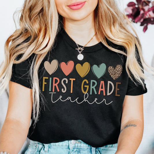 5 hearts in a row, all different colors and one cheetah print. "first grade teacher" underneath the hearts, "ifrst grade" text is each different colors. "teacher" in cursive underneath. Women Unisex Tshirt Color: Black