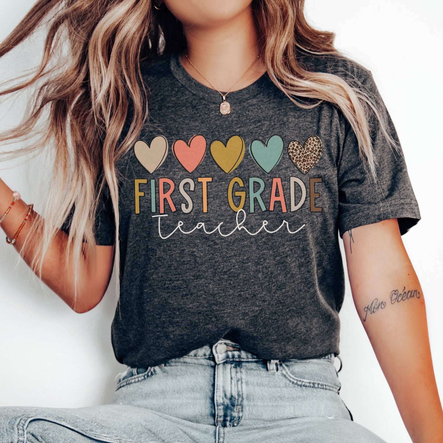 5 hearts in a row, all different colors and one cheetah print. "first grade teacher" underneath the hearts, "ifrst grade" text is each different colors. "teacher" in cursive underneath. Women Unisex Tshirt Color: Dark Grey Heather