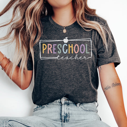 Preschool Teacher Tshirt Multicolor font with white rectangle borders and an apple on the top. Dark Grey Heather color