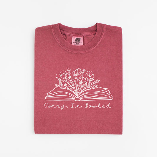 A folded unisex Comfort Colors t-shirt in crimson color. The shirt features a line drawing of an open book with flowers and plants growing from it, accompanied by the text 'Sorry, I'm booked' below the design.