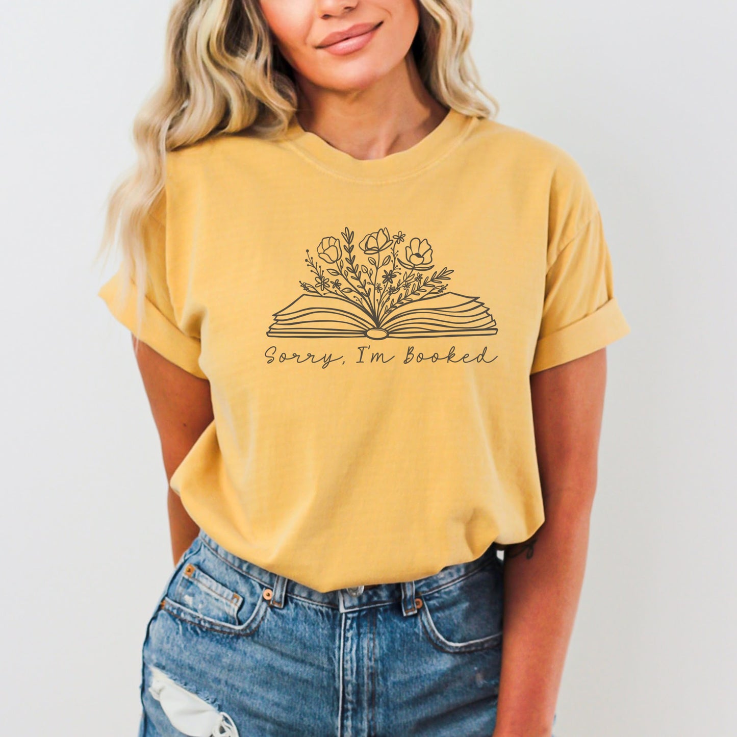 A woman wearing a unisex Comfort Colors t-shirt in mustard color. The shirt features a line drawing of an open book with flowers and plants growing from it, with the text 'Sorry, I'm booked' below the design.