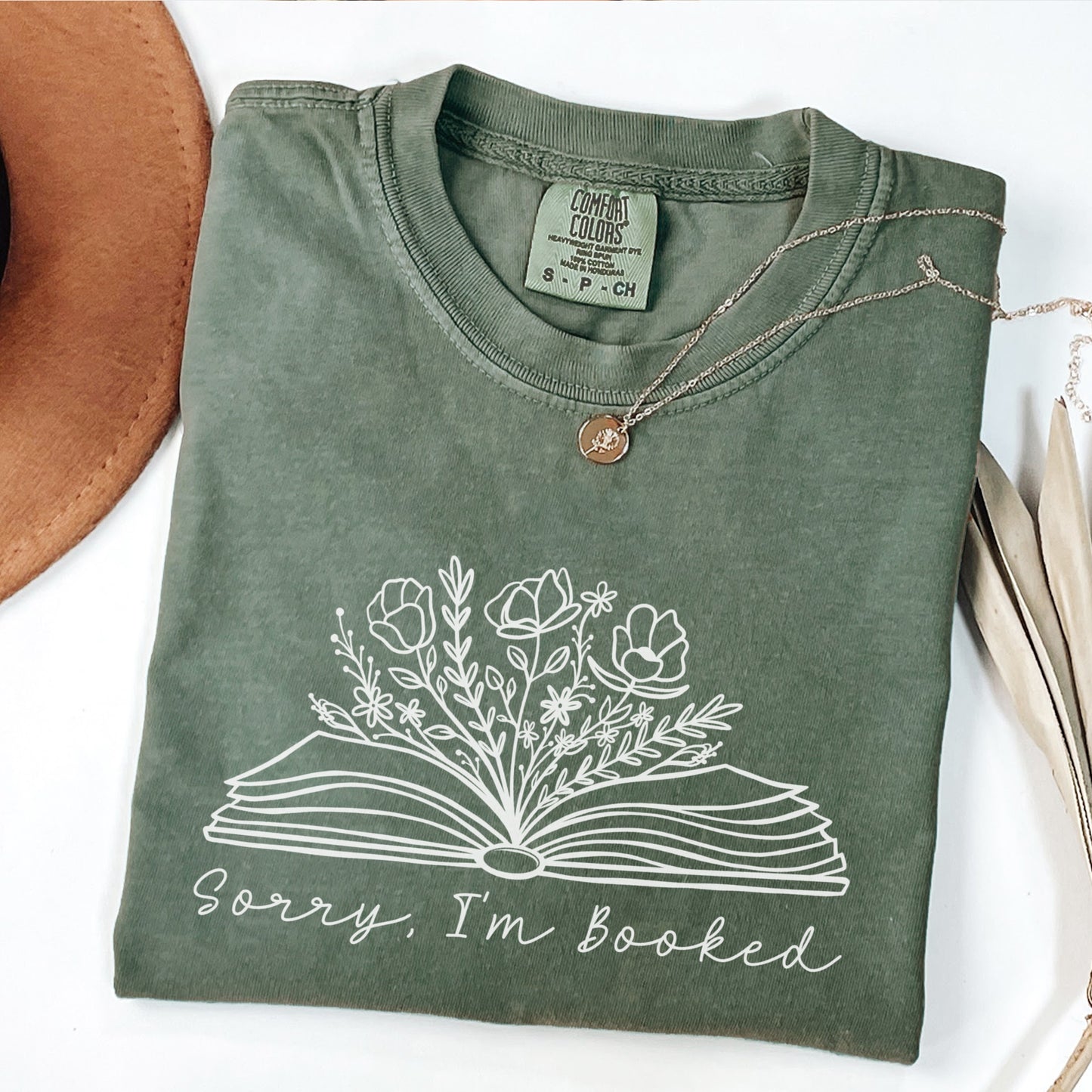 A folded unisex Comfort Colors t-shirt in moss color, displayed with a necklace and a hat beside it. The shirt features a line drawing of an open book with flowers and plants growing from it, with the text 'Sorry, I'm booked' below the design.