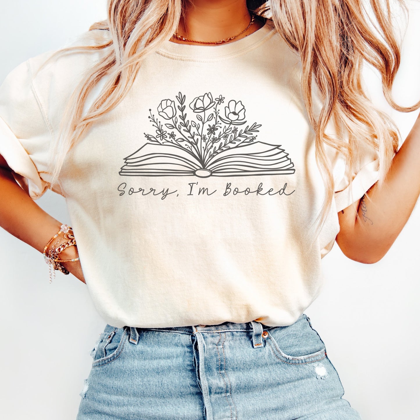 A woman wearing a unisex Comfort Colors t-shirt in ivory color. The shirt displays a line drawing of an open book with flowers and plants growing from it, with the text 'Sorry, I'm booked' below the design.
