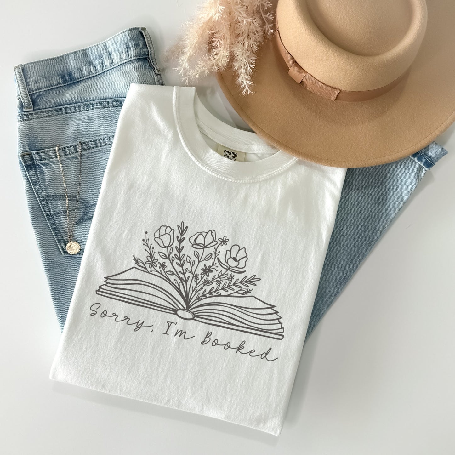 A folded unisex Comfort Colors t-shirt in white, displayed with a pair of light blue jeans, a tan hat, and a necklace. The shirt features a line drawing of an open book with flowers and plants growing from it, with the text 'Sorry, I'm booked' below the design.