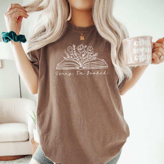 Person wearing an espresso-colored unisex Comfort Colors t-shirt with the phrase 'Sorry, I’m Booked' in white script above an open book graphic with flowers sprouting from the pages. The person holds a mug and has long blonde hair, creating a cozy book lover aesthetic.