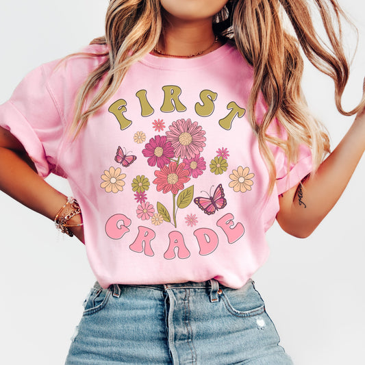 "First Grade" with 'first' in tan font, 'grade' in pink font. 'first' above a design of wildflowers and butterflies. 'Grade' underneath. Adult Unisex Tshirt Color: Blossom