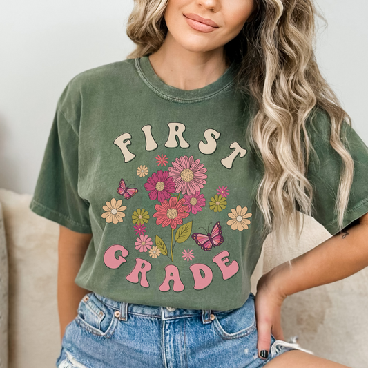 "First Grade" with 'first' in tan font, 'grade' in pink font. 'first' above a design of wildflowers and butterflies. 'Grade' underneath. Adult Unisex Tshirt Color: Moss