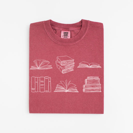 A folded unisex Comfort Colors t-shirt in crimson color. The shirt features white line drawings of various book illustrations, including open and stacked books.