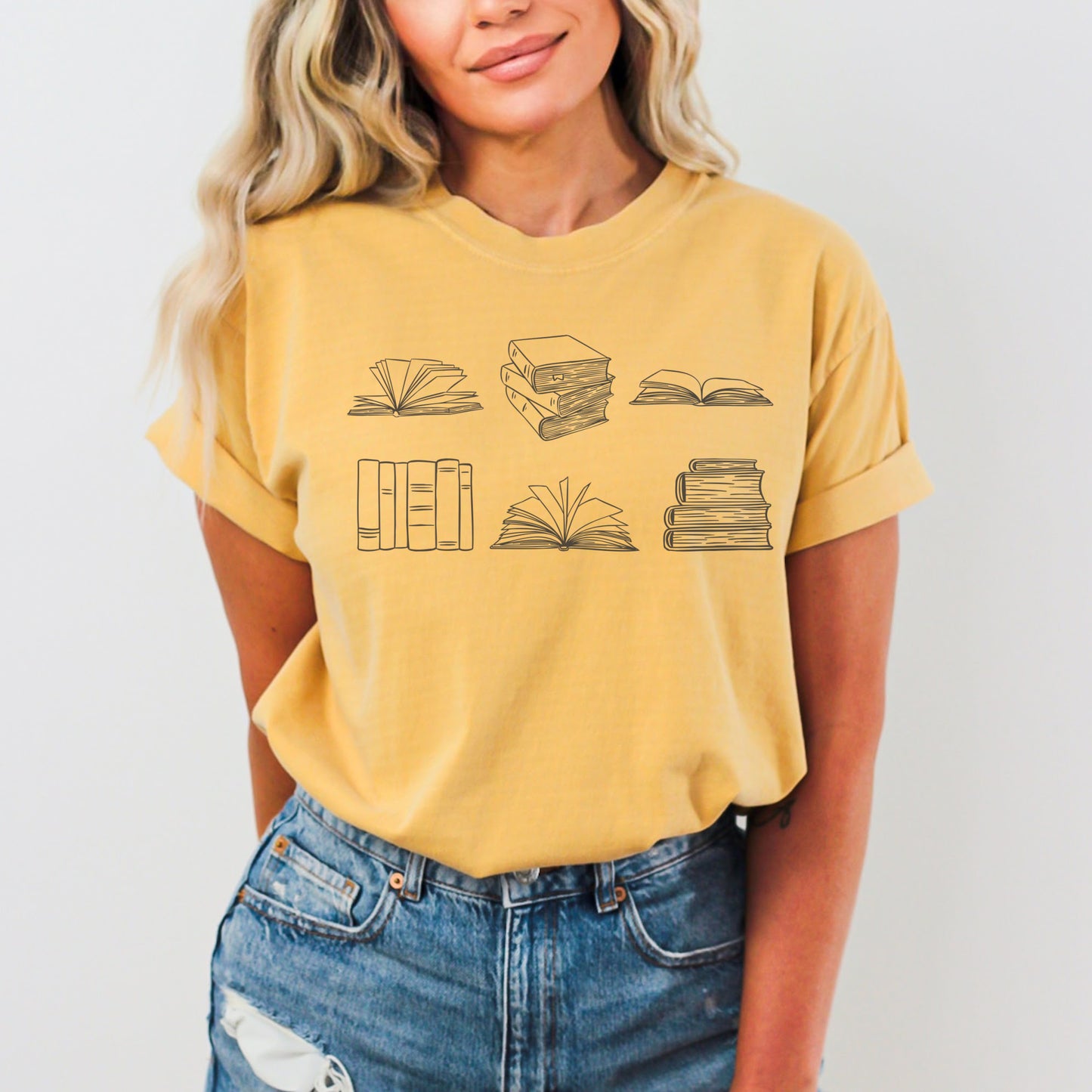 A woman wearing a unisex Comfort Colors t-shirt in mustard color. The shirt features brown line drawings of various book illustrations, including open and stacked books.