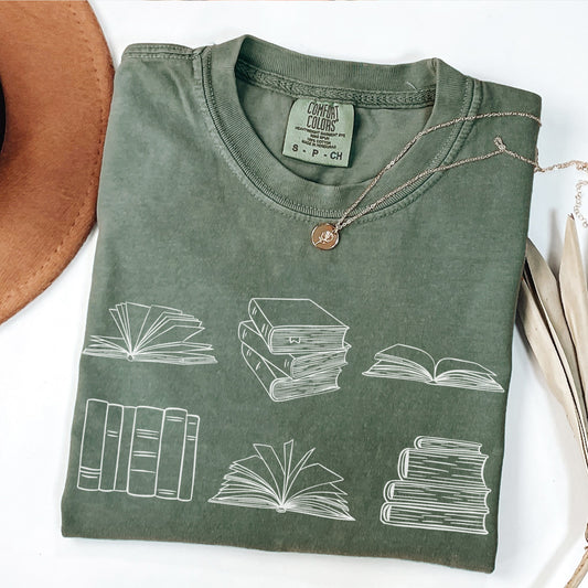 A folded unisex Comfort Colors t-shirt in moss color, displayed with a necklace and a hat beside it. The shirt features white  line drawings of various book illustrations, including open and stacked books.