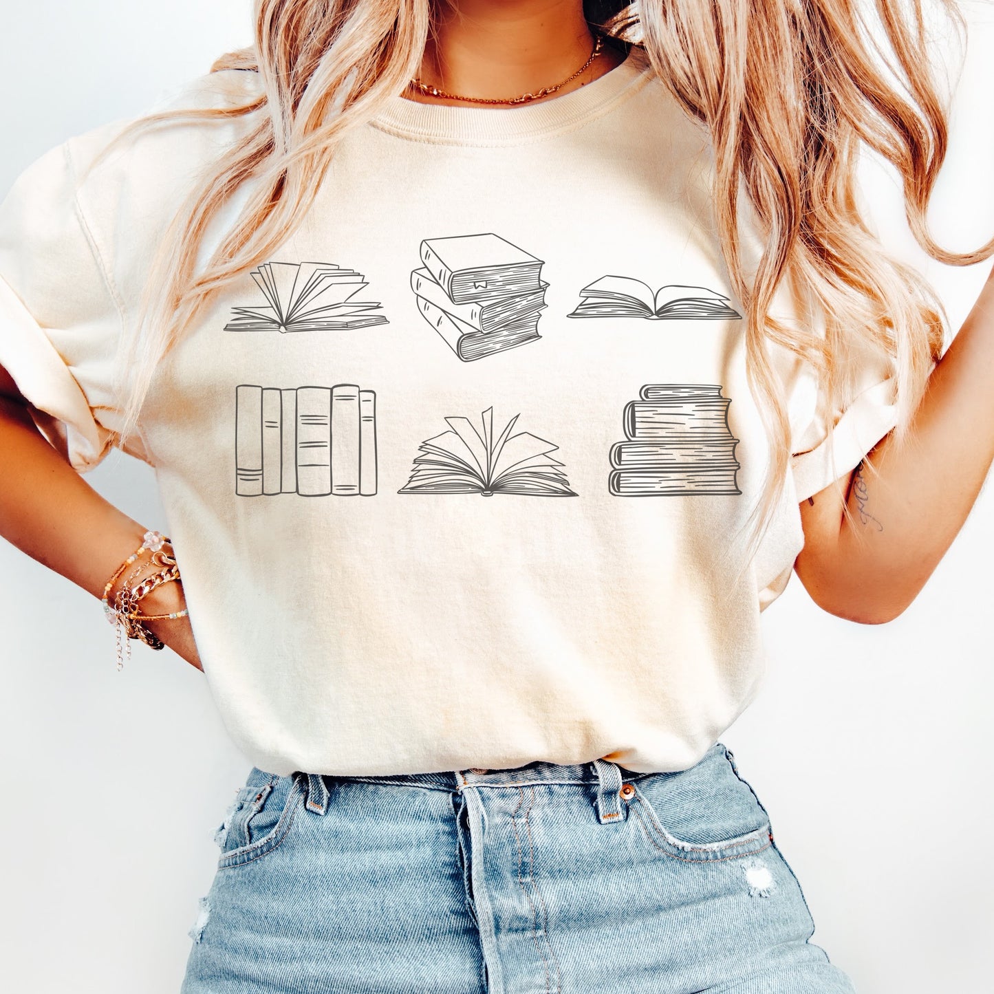 A woman wearing a unisex Comfort Colors t-shirt in ivory color. The shirt features brown line drawings of various book illustrations, including open and stacked books.