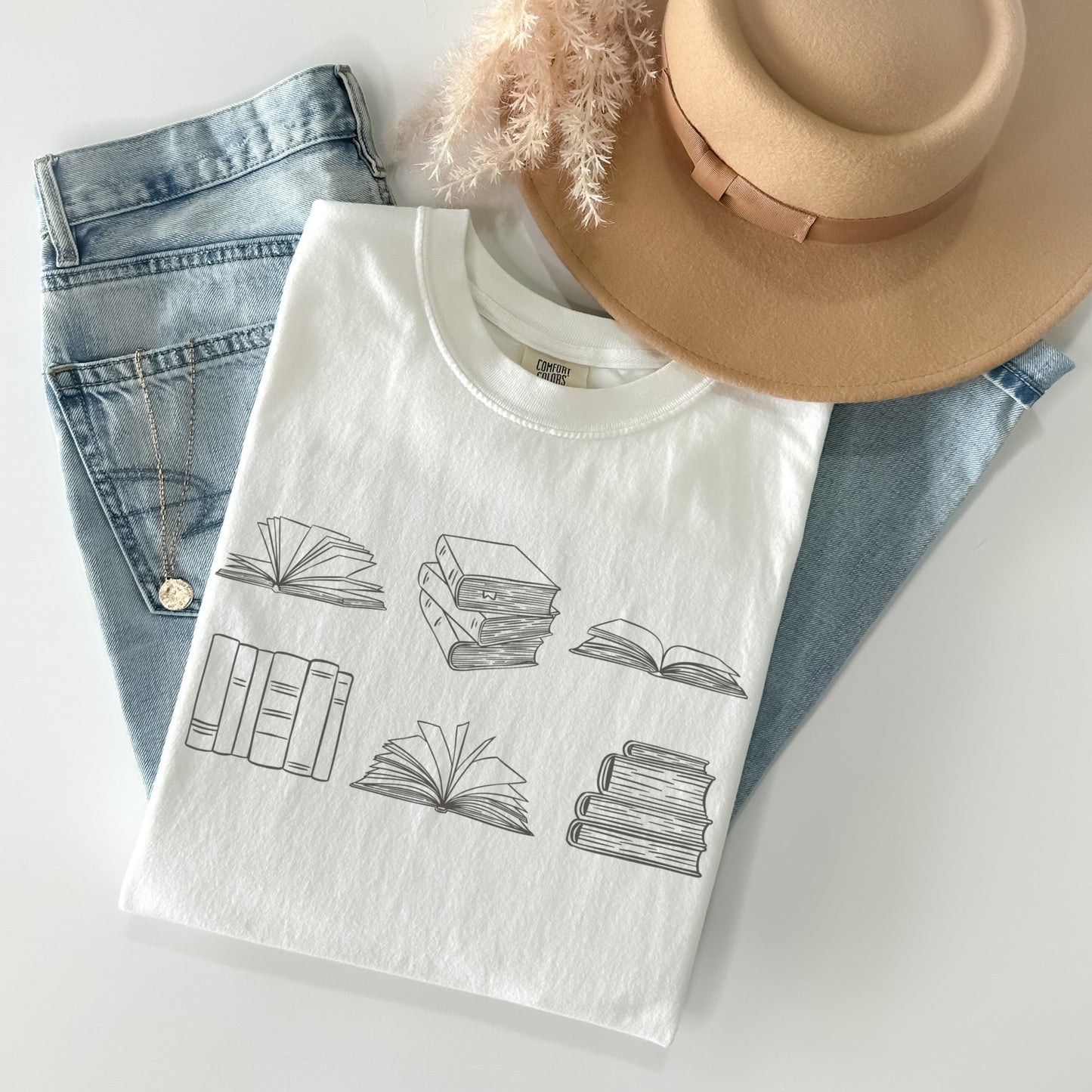 A folded unisex Comfort Colors t-shirt in white, displayed with a pair of light blue jeans, a tan hat, and a necklace. The shirt features brown line drawings of various book illustrations, including open and stacked books.