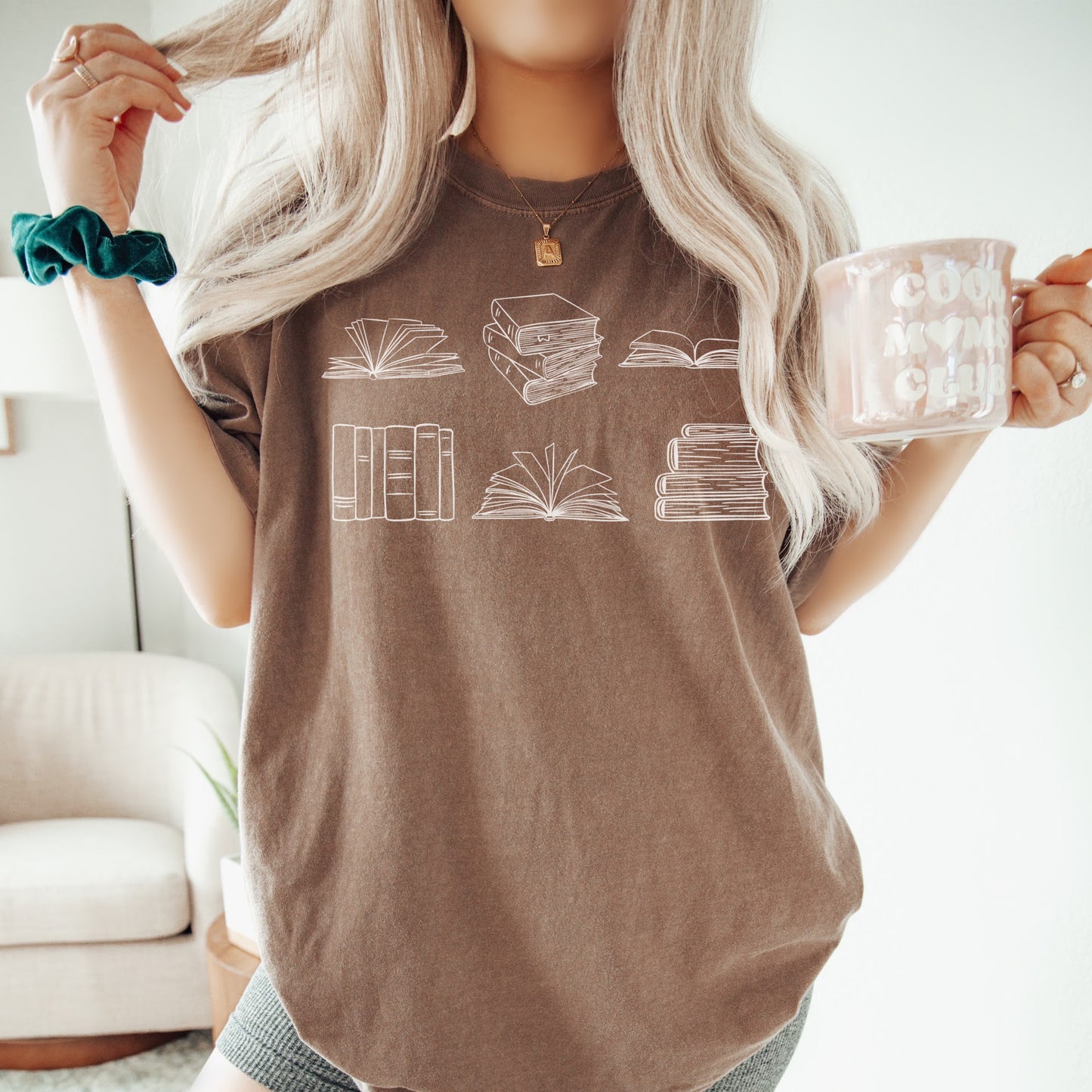 A woman wearing a unisex Comfort Colors t-shirt in espresso color. The shirt features white line drawings of various book illustrations, including open and stacked books.