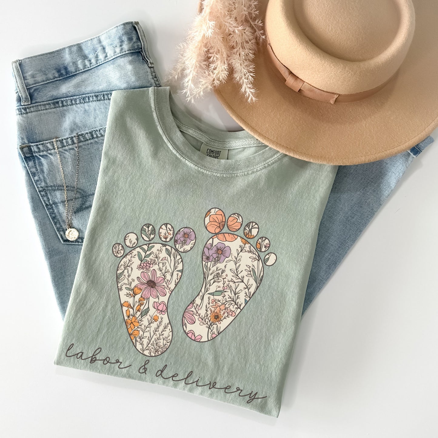 Tshirt with a graphic of two baby feet with floral design. "Labor and Delivery" in cursive underneath the footprints. Shirt Color: Bay