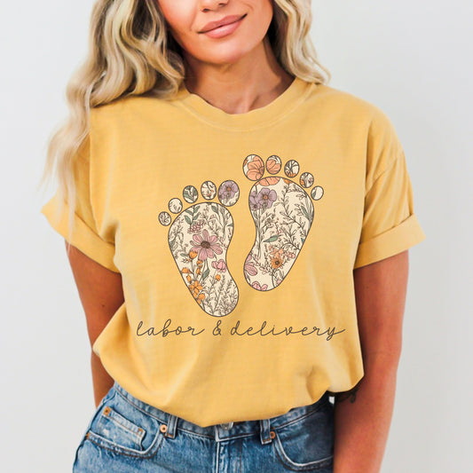 Tshirt with a graphic of two baby feet with floral design. "Labor and Delivery" in cursive underneath the footprints. Shirt Color: Mustard