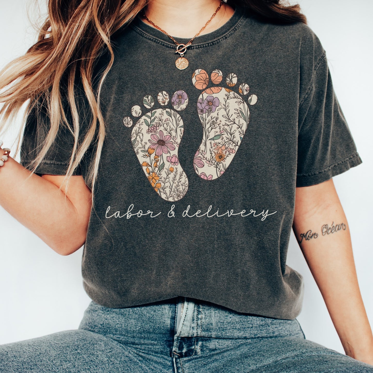 Tshirt with a graphic of two baby feet with floral design. "Labor and Delivery" in cursive underneath the footprints. Shirt Color: Pepper