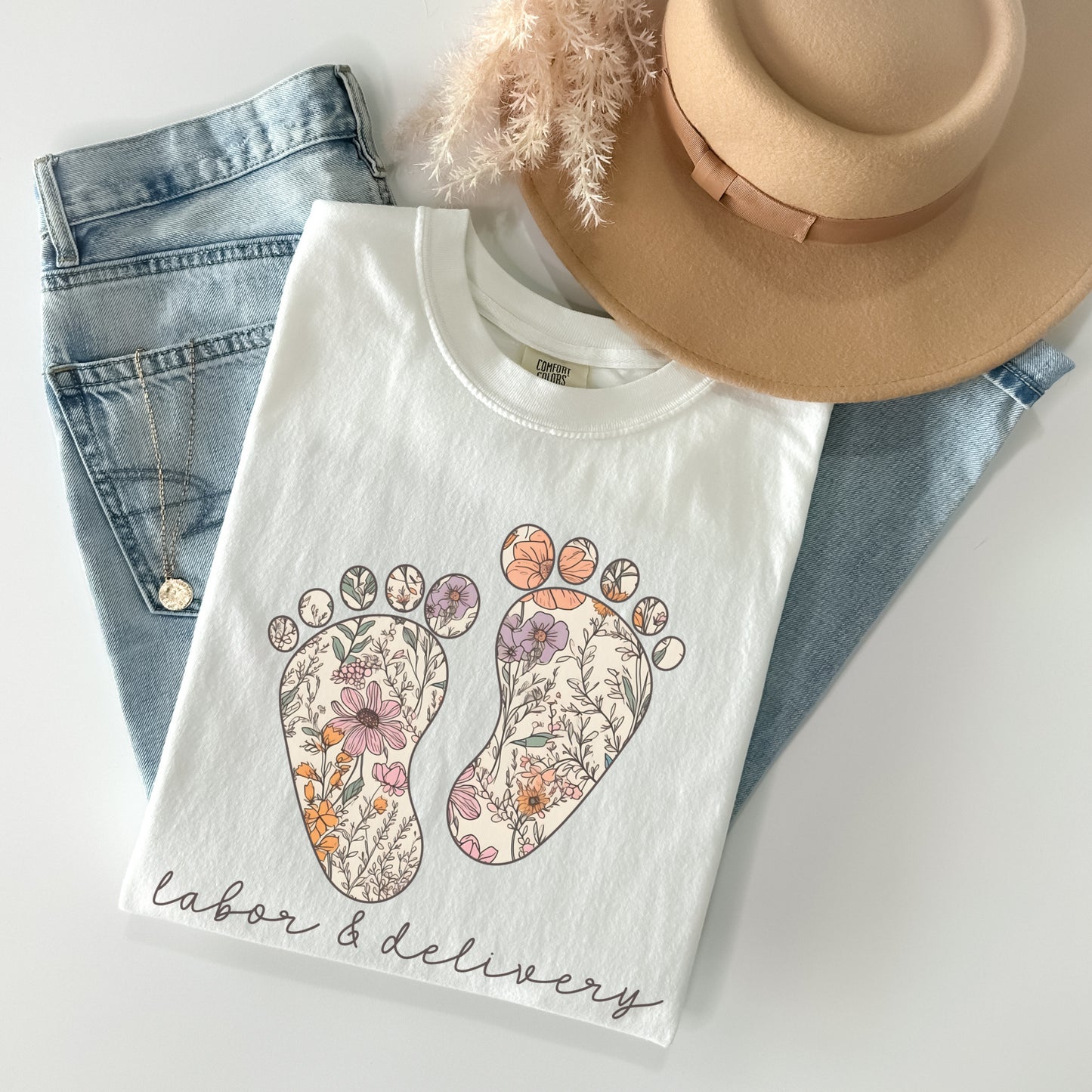 Tshirt with a graphic of two baby feet with floral design. "Labor and Delivery" in cursive underneath the footprints. Shirt Color: White