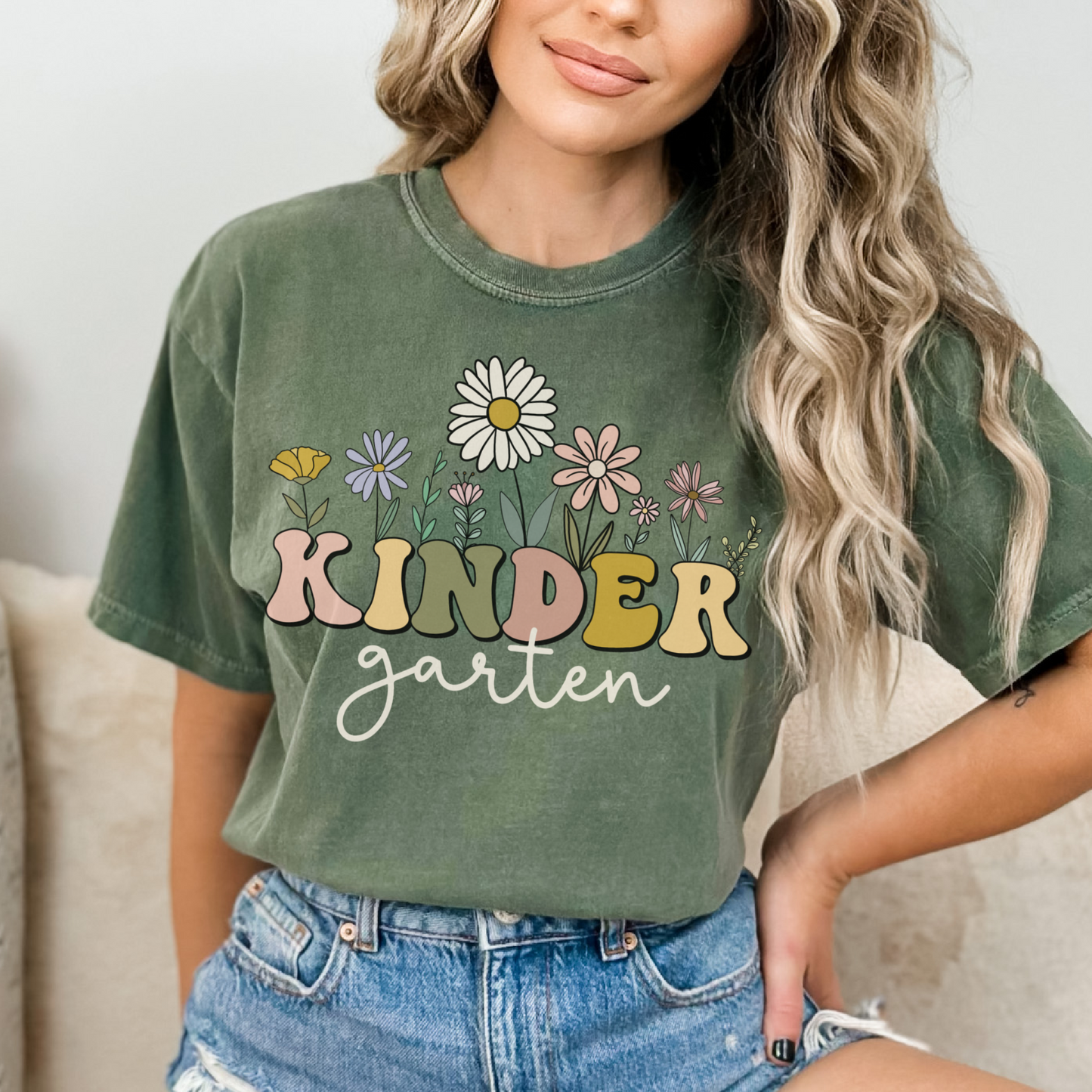 Comfort Colors® Tshirt that says "Kindergarten". 'KINDER' is in a bold, multicolored font with wildflowers coming out of the top of it. 'garten' is underneath in a light cursive font. Tshirt color: Moss