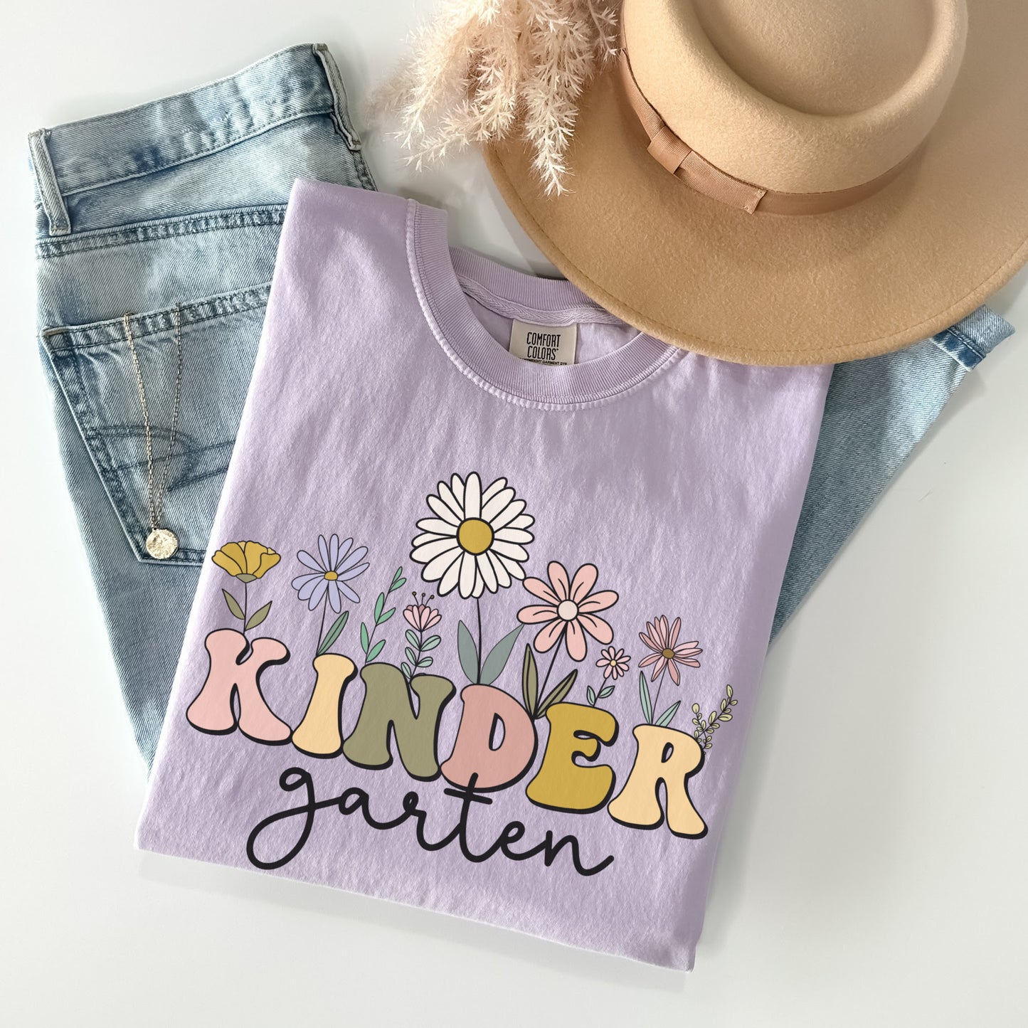 Comfort Colors® Tshirt that says "Kindergarten". 'KINDER' is in a bold, multicolored font with wildflowers coming out of the top of it. 'garten' is underneath in a black cursive font. Tshirt color: Orchid