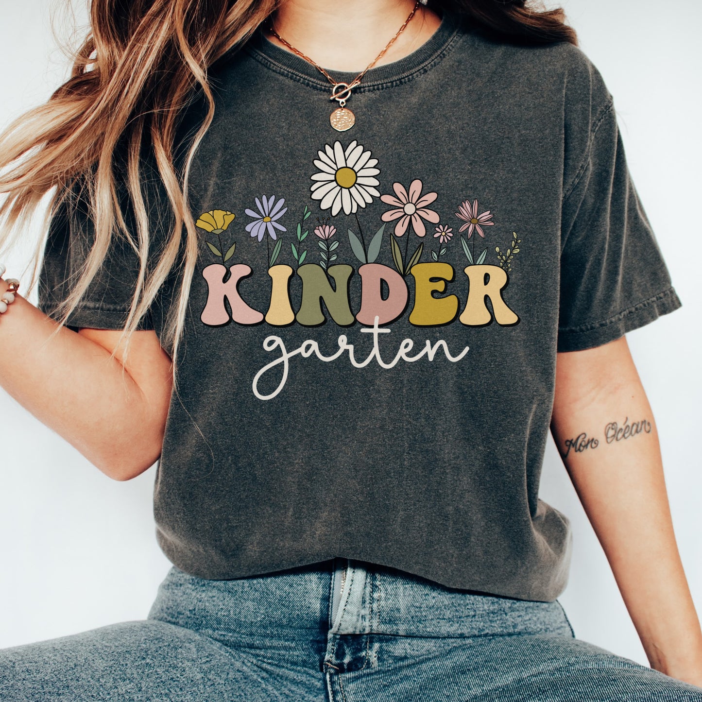 Comfort Colors® Tshirt that says "Kindergarten". 'KINDER' is in a bold, multicolored font with wildflowers coming out of the top of it. 'garten' is underneath in a light cursive font. Tshirt color: Pepper