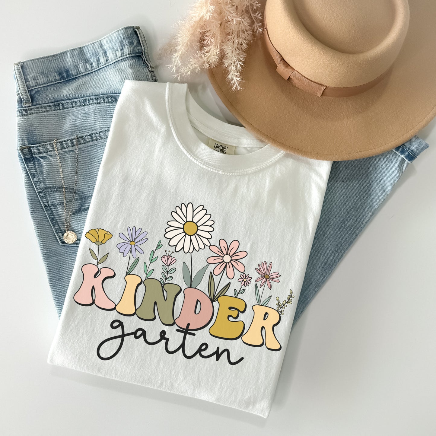 Comfort Colors® Tshirt that says "Kindergarten". 'KINDER' is in a bold, multicolored font with wildflowers coming out of the top of it. 'garten' is underneath in a black cursive font. Tshirt color: