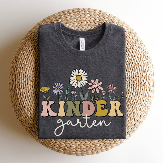 Tshirt that says "Kindergarten". 'KINDER' is in a bold, multicolored font with wildflowers coming out of the top of it. 'garten' is underneath in a light cursive font. Tshirt color: Dark Grey Heather