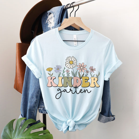 Tshirt that says "Kindergarten". 'KINDER' is in a bold, multicolored font with wildflowers coming out of the top of it. 'garten' is underneath in a black cursive font. Tshirt color: Heather Ice Blue