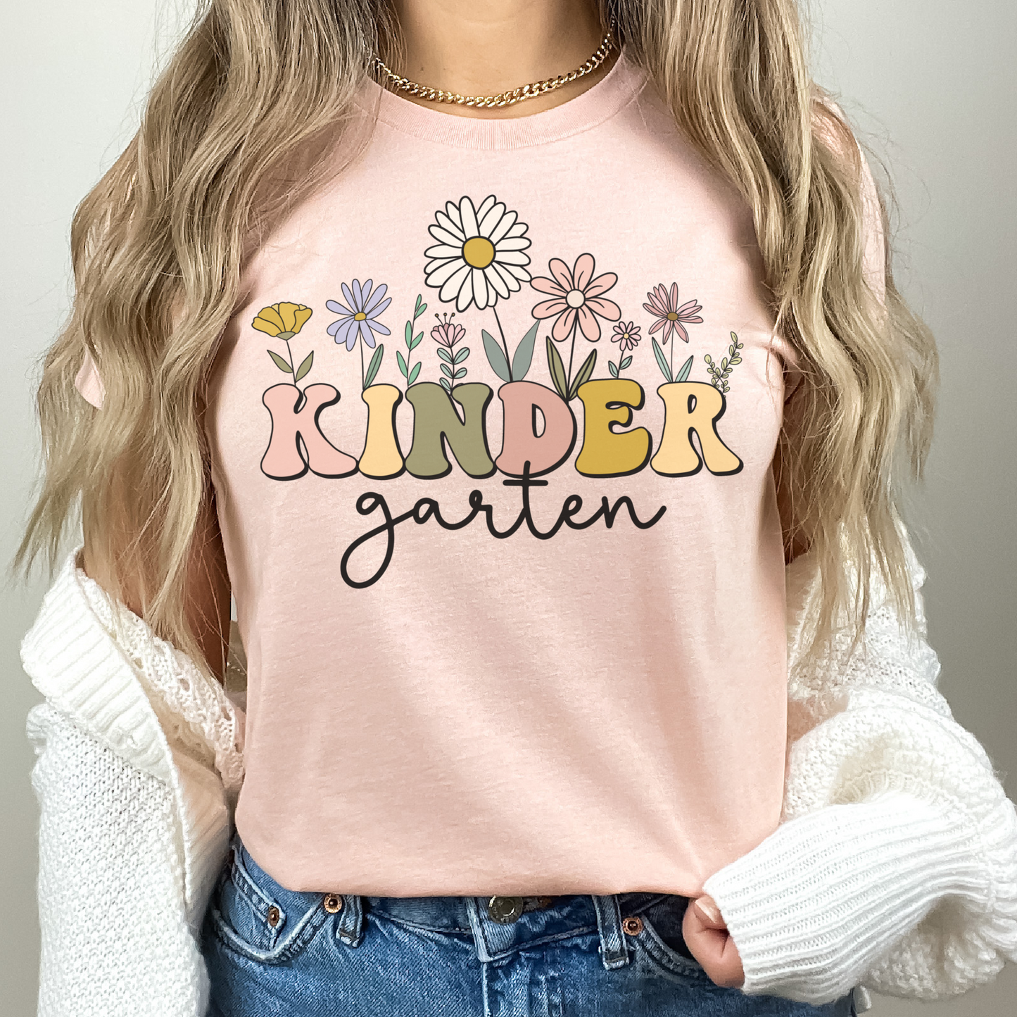 Tshirt that says "Kindergarten". 'KINDER' is in a bold, multicolored font with wildflowers coming out of the top of it. 'garten' is underneath in a black cursive font. Tshirt color: Heather Peach