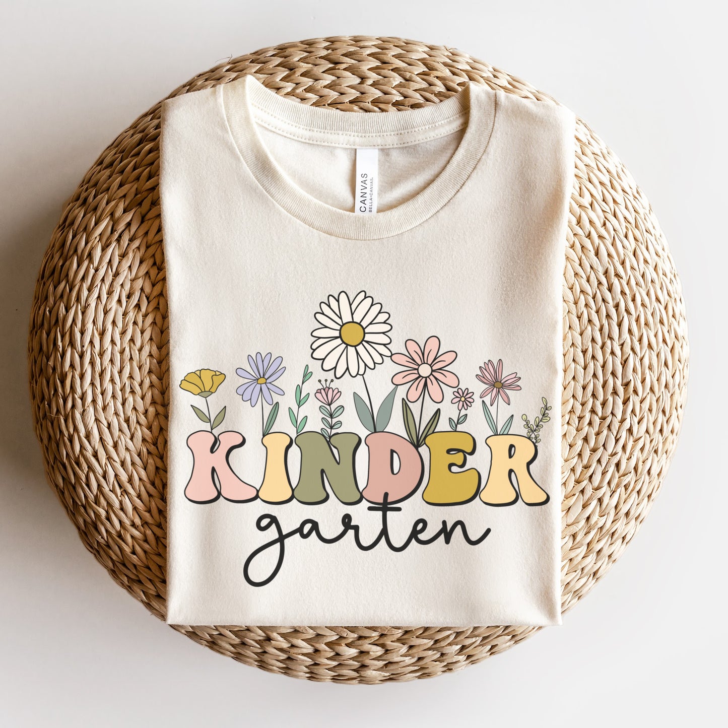 Tshirt that says "Kindergarten". 'KINDER' is in a bold, multicolored font with wildflowers coming out of the top of it. 'garten' is underneath in a black cursive font. Tshirt color: Natural