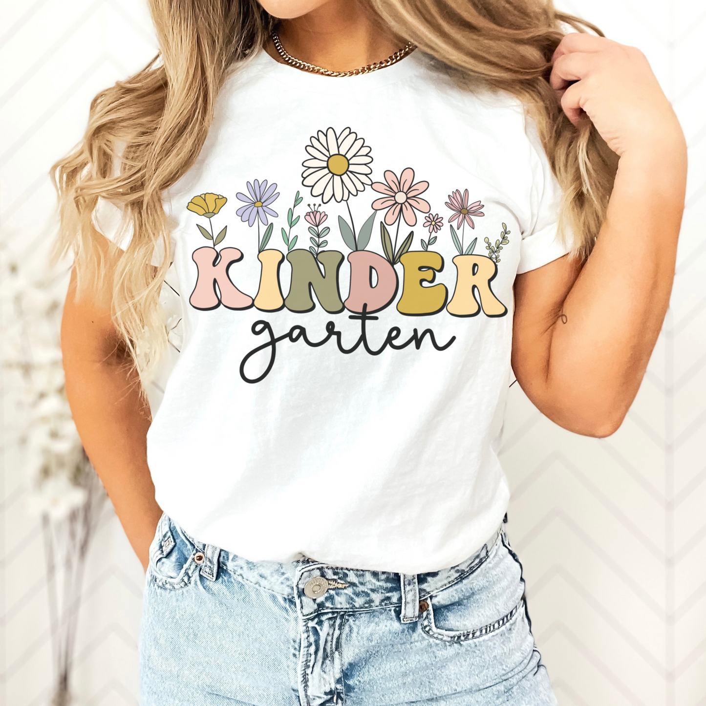 Tshirt that says "Kindergarten". 'KINDER' is in a bold, multicolored font with wildflowers coming out of the top of it. 'garten' is underneath in a black cursive font. Tshirt color: White